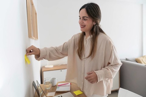 Smiling content creator placing yellow sticky note on wall next to framed print, desk with laptop