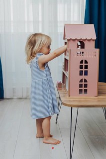 Child in a blue dress exploring a pink dollhouse on a wooden table in a brightly lit room