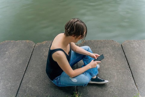 Woman Using Smartphone Sitting by the Water's Edge, A person sits on a concrete surface by the