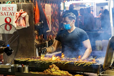 Street food vendor turns and glazes satay skewers over open flames at Satay Street street-food