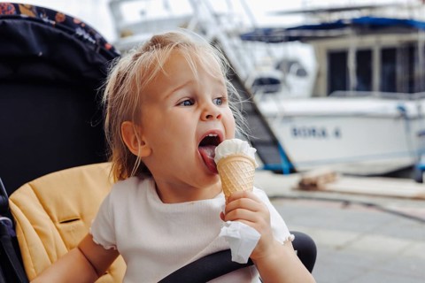 A young two year girl relishes a cone of white ice cream while visiting a port with boats on a