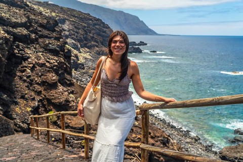 Young woman smiling while standing on a path with a wooden handrail, overlooking the deep blue