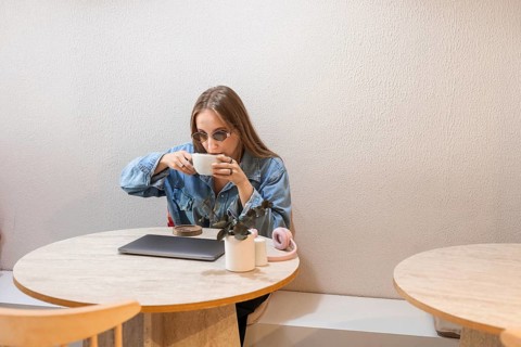 A young Caucasian woman with long brown hair sits at a wooden table, sipping coffee from a white