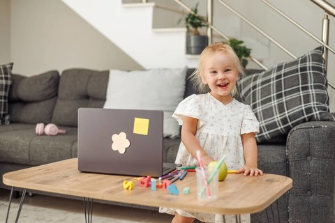 Blond toddler girl standing at wooden table with colorful toys and laptop in modern living room