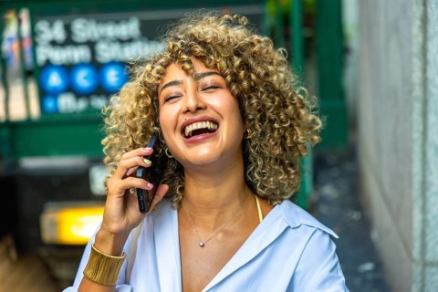 Happy young woman with curly hair laughing while having a phone call, standing in front of a