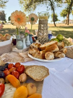 A picnic spread featuring fresh fruits, bread, and pastries on a white tablecloth. Flowers and