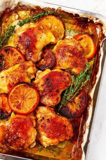 A tray holds cooked chicken thighs topped with orange slices and fresh herbs. The chicken has a