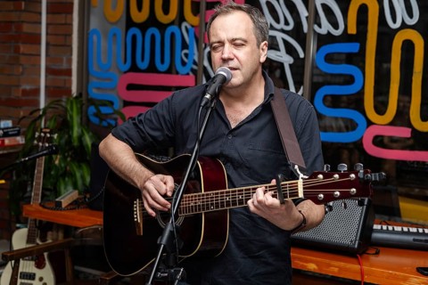 A middle-aged Caucasian man with short brown hair sings and plays an acoustic guitar in a vibrant