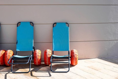 Two blue beach loungers with orange wheels are positioned against a gray wall. The loungers are