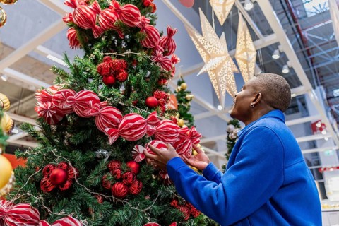 African man decorates a Christmas tree with red ornaments and ribbons in a festive indoor setting.
