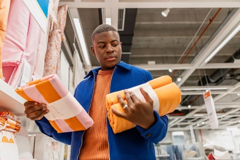 A young Black man examines colorful towels in a retail store. He holds an orange towel in one hand