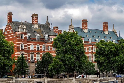 Mature riverside trees frame the ornate red brick facade of the Norman Shaw Building on Victoria