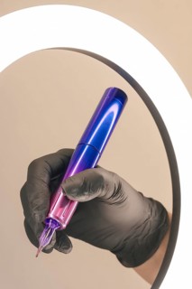 A gloved hand holding a purple and blue tattoo machine in front of an oval mirror
