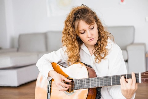 Focused woman playing guitar on a sofa, wearing a white shirt
