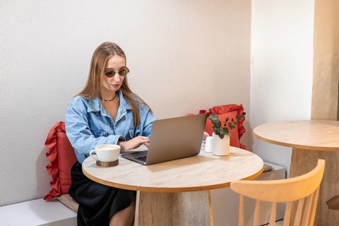 Young Caucasian woman with long brown hair wearing sunglasses, working on a laptop at a table with