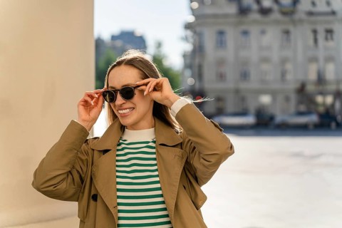 Smiling woman puts on sunglasses outside in the city, A woman in a trench coat and sunglasses