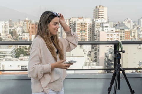 Smiling woman lifting sunglasses off her head, holding smartphone near tripod, city skyline