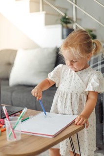 Young blond European girl drawing with colored pencils at a table in a living room with a laptop