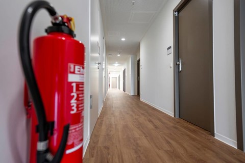 Modern corridor with light wooden floors, fire extinguisher in the foreground and several doors,