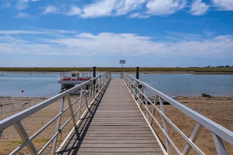 Wooden walkway leading to water with boat on shore and blue sky, Santa Luzia, Tavira, Faro,