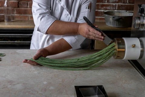 A chef skillfully extrudes vibrant green noodles from a pasta machine in a modern kitchen. The