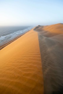 Sand dunes with wave patterns and sea in evening light, wind blowing sand across the dunes,