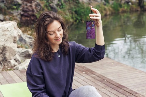 Young Caucasian woman with brown hair sitting by a lake, holding a smartphone. She wears a dark