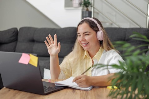 Young blond European woman with headphones waving hand during video call at home with laptop and