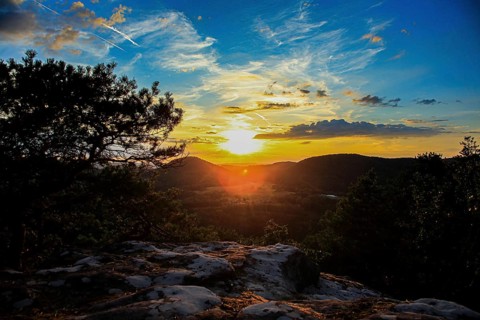 Sunset over wooded hills with clouds in the sky. Rocks in the foreground create a quiet, peaceful