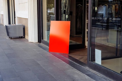 Mockup, vibrant red sign leans against entrance of clothing store. Midday sun casts light on modern