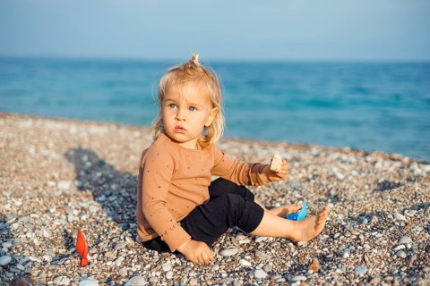 A young child sits on a pebble beach, holding a snack in one hand. The child girl appears curious