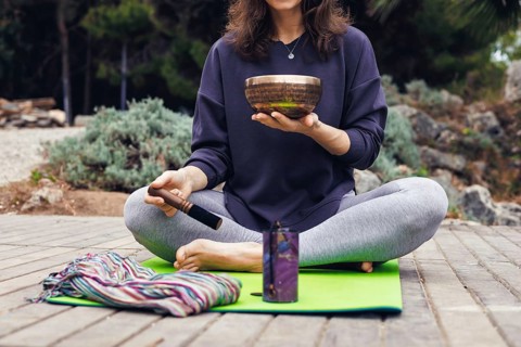 A young Caucasian woman with long brown hair sits cross-legged on a green yoga mat by a lake. She