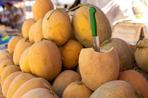 Ripe melons stacked at an outdoor market with one melon cut open displaying its flesh