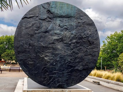 A weathered stone disc with a rippled, wind carved surface stands on its own plinth in the gardens