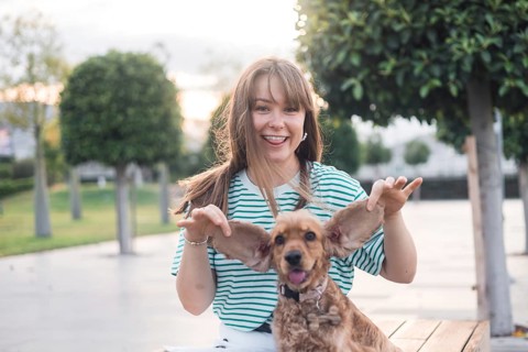 A woman laughs while playfully posing with her dog on a bench in a park