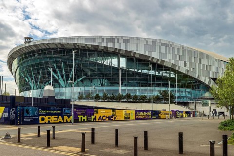 The curved glass and metal facade of Tottenham Hotspur Stadium rises above hoarding on White Hart