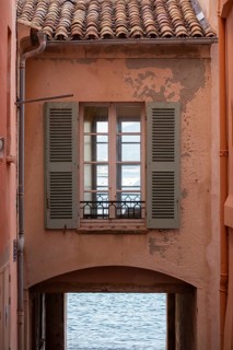 A shuttered window on a weathered ochre facade sits above an arched passage opening toward the sea,