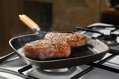 Two beef steaks are cooking in cast iron skillet on stovetop in kitchen. Smoke rises from the