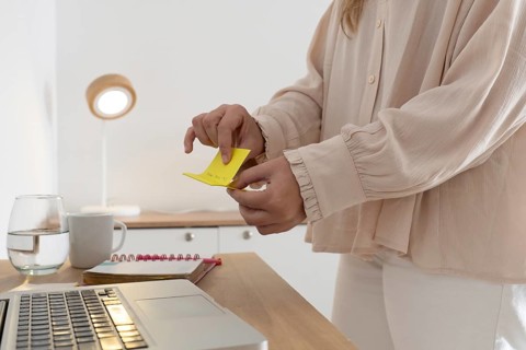Detailed close up of woman's hands holding and reading a yellow sticky note reminder near laptop,