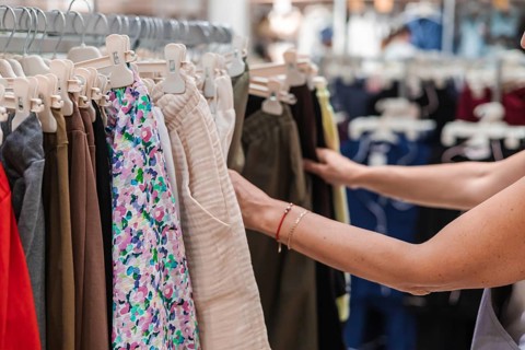 A woman with light skin and brown hair selects clothing from a rack in a retail store. The scene