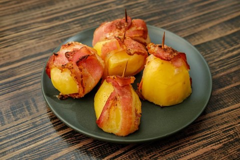 Plate with roasted potatoes wrapped in bacon on wooden table. Slices of bacon secured with