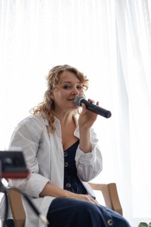 A young Caucasian woman with curly hair sings into a microphone at a home concert. Natural light