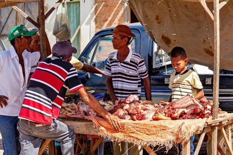 Group of people discussing meat products at a market stand, interacting under tent tarpaulins,