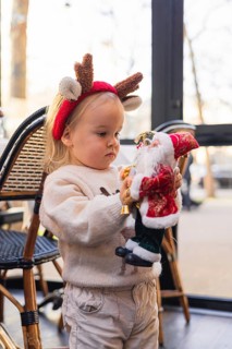 A young Caucasian girl with blonde hair wears reindeer antlers. She holds a Santa Claus figurine,