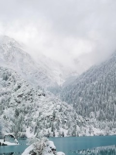 Ice-covered turquoise water of a mountain lake against the backdrop of mountains in fog and clouds.