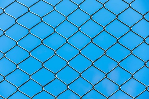 A close-up view of a chain-link fence against a clear blue sky. The fence features a diamond
