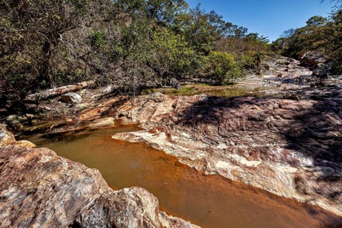 A dry riverbed with sparse water flow between rocky landscape and trees, a small river flows