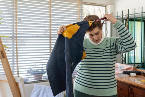 Older woman lifts a blue denim apron with yellow detail over her head to remove it, standing near a