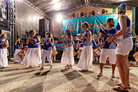 Women in white skirts and blue tops dance to drum music at a nighttime show, a band plays and
