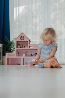 Child toddler blonde girl in blue dress playing with a wooden pink toy house at home